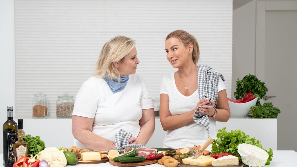 Two women in a kitchen preparing fresh vegetables and cheese with olive oil and spices.